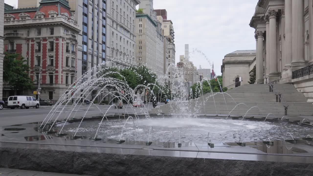 The video dollys between slender spring trees leading the viewer towards an artistically crafted fountain located in front of New York City's
