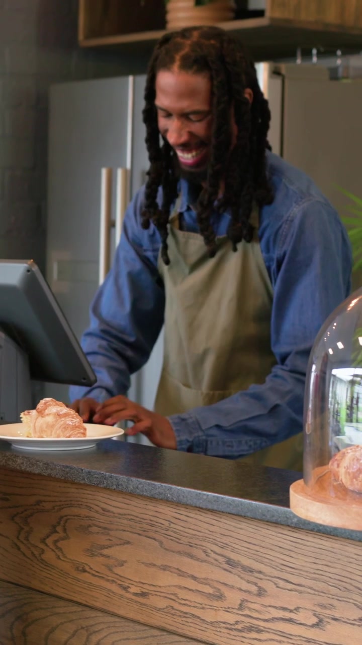 Vertical video: Tapping POS, server at cafe counter in green apron serving guest, nudging croissant