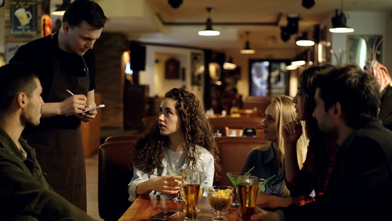 Young people are talking with waiter making order in cafe while waiter is listening to customers and writing in notepad with pen. Eating and drinking out concept.
