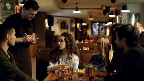 young people are talking with waiter making order in cafe while waiter is listening to customers and writing in notepad with pen. eating and drinking out concept.