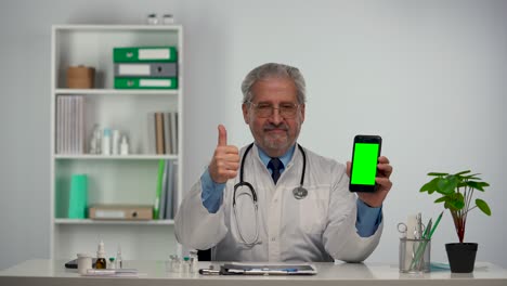 aged male doctor holds smartphone with green screen chroma key, points at it with finger and makes thumb up gesture. doc in white medical coat sits at table in hospital office. slow motion