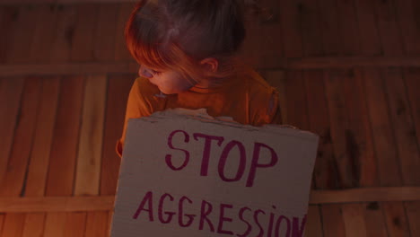 Afraid-homeless-toddler-girl-sitting-holding-inscription-Stop-Aggression,-hiding-from-bombing-attack