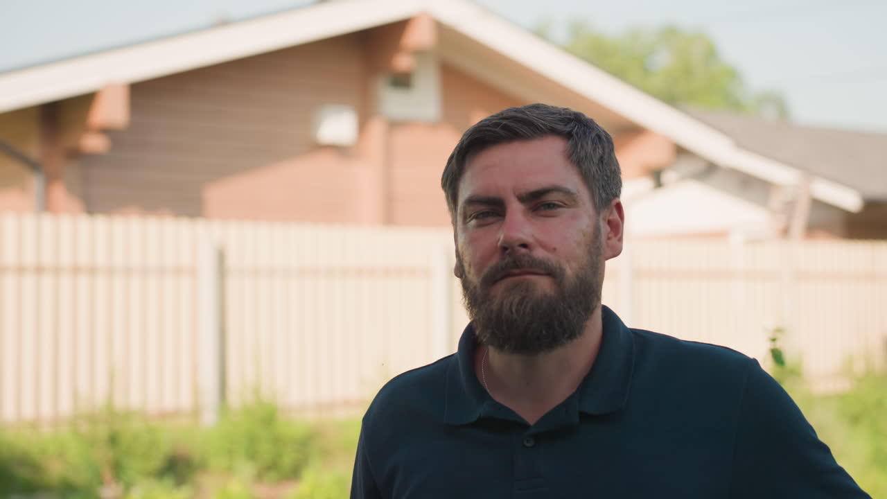 Bearded man in dark polo outdoors with hand on waist turns and gazes sideways near wooden house and fence, sunny backyard, calm thoughtful expression, rural setting