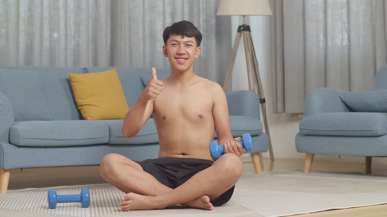 집에서 셔츠를 벗고 운동하는 동안 매트에 앉아서 웃고 엄지손가락을 올리는 아시아 십대 소년