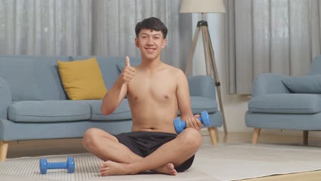 asian teenager boy with dumbbells smiling and showing thumbs up gesture while sitting on mat during the shirtless workout at home