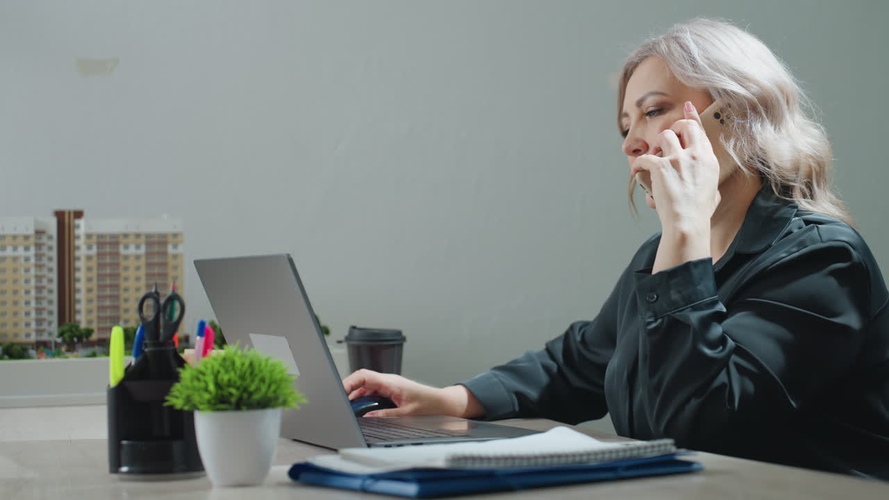 Real estate agent retrieves phone from folder, makes business call while reviewing property model displayed on desk and browsing listings on laptop, focused on multitasking in office setting