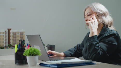 real estate agent retrieves phone from folder, makes business call while reviewing property model displayed on desk and browsing listings on laptop, focused on multitasking in office setting