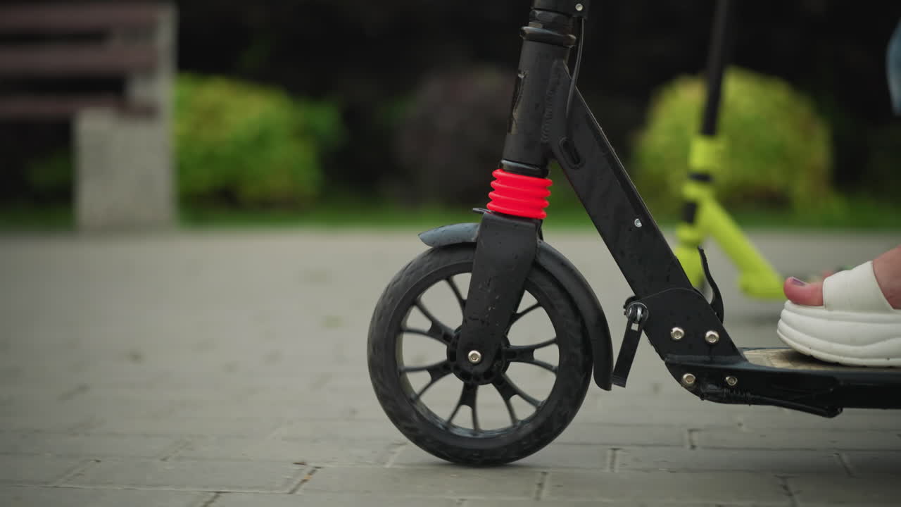 Close-up leg view of a woman riding a black scooter, her legs visible as she moves forward, a small child follows behind on a green scooter