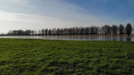 Flying-Over-Green-Grass-Towards-Frozen-River-At-Sandelingen-Park-In-Hendrik-Ido-Ambacht,-Netherlands