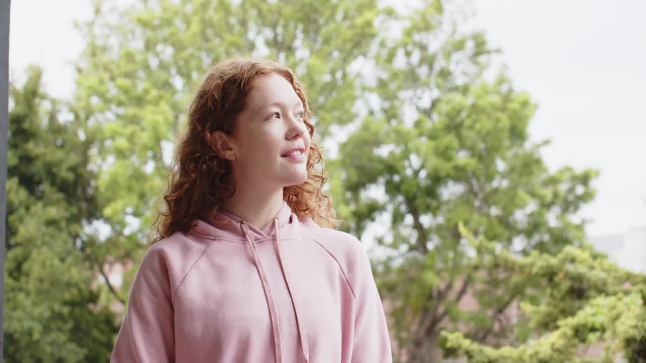 Donna adulta che nota la fotocamera, si gira verso l'obiettivo, sorridendo su un portico con felpa rosa e alberi
