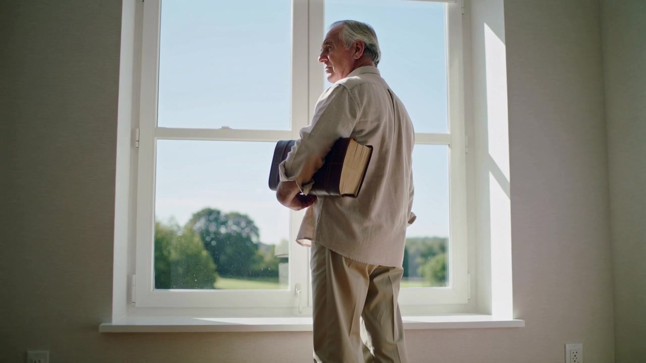 Sun through window as senior at home shifting, holding book and jacket, viewing lawn, beige top