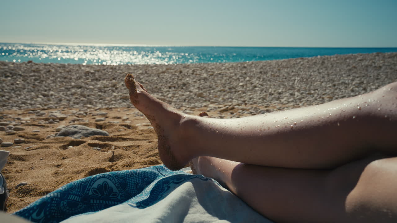 Sunlit legs rest on a beach towel beside warm sand and pebbles, facing a sparkling sea that suggests summer ease, leisure, and carefree coastal escape