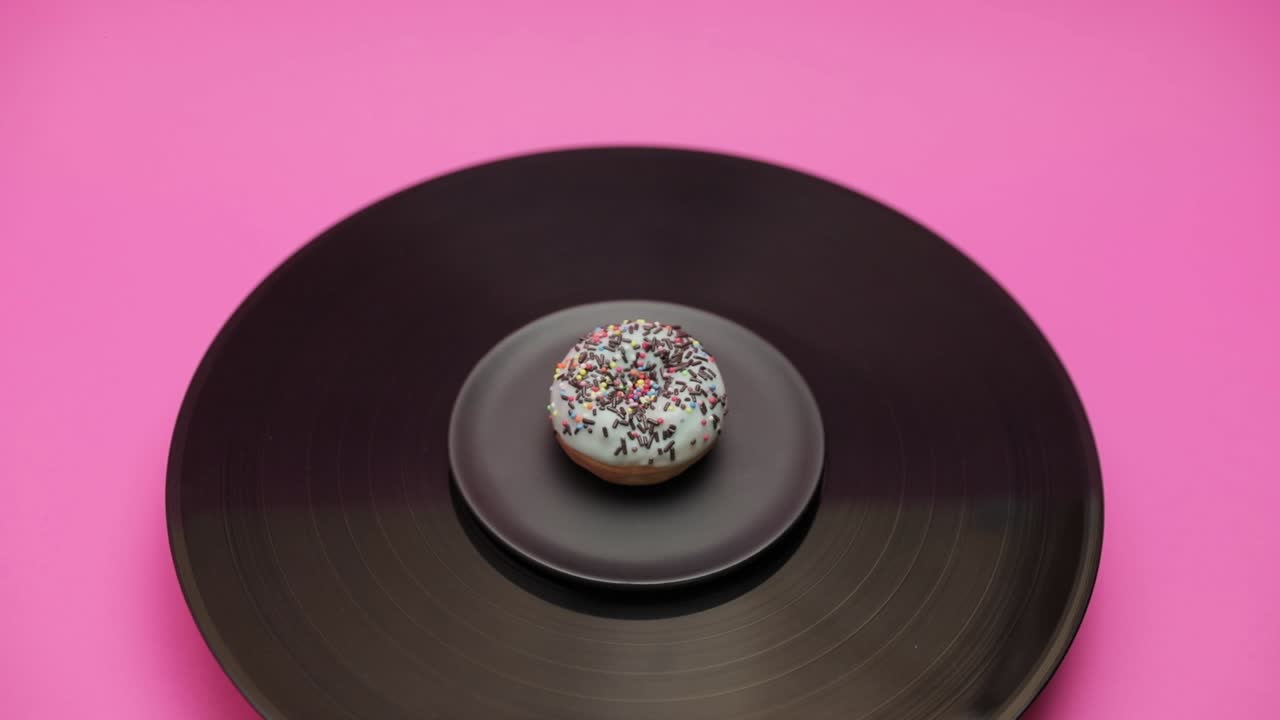 An American donut topped with blue chocolate with black choco and colorful crumbs turns in a circle on a black turntable. The shot from above