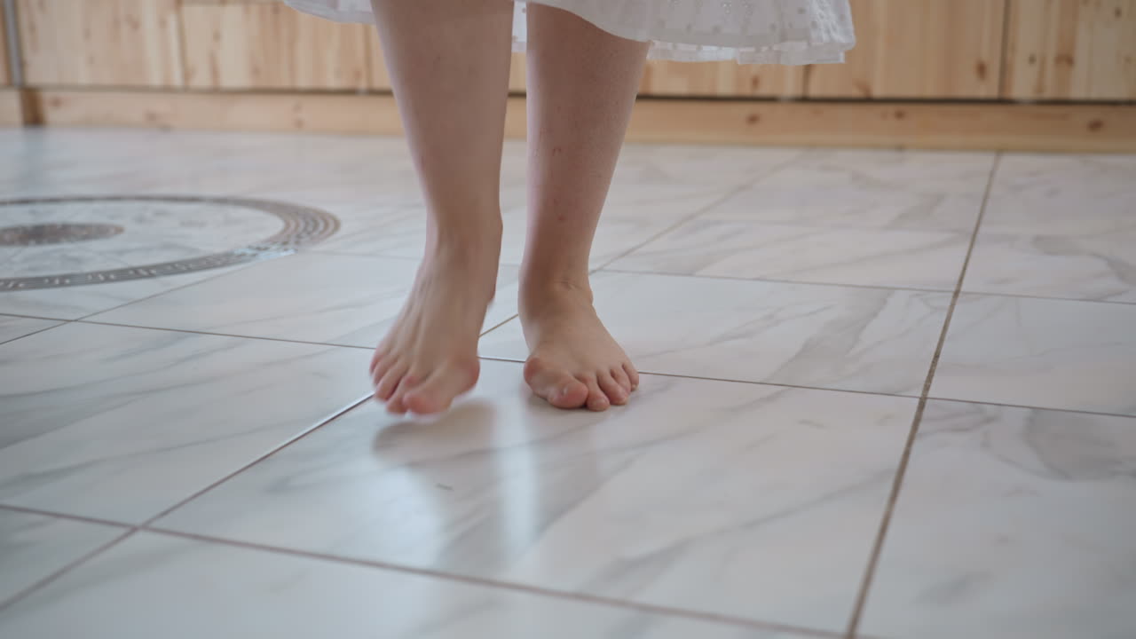 Woman tiptoeing across glossy marble floor toward round glass table in sunlit kitchen, barefoot silent steps showing careful balance in calm domestic scene with subtle rustic warmth