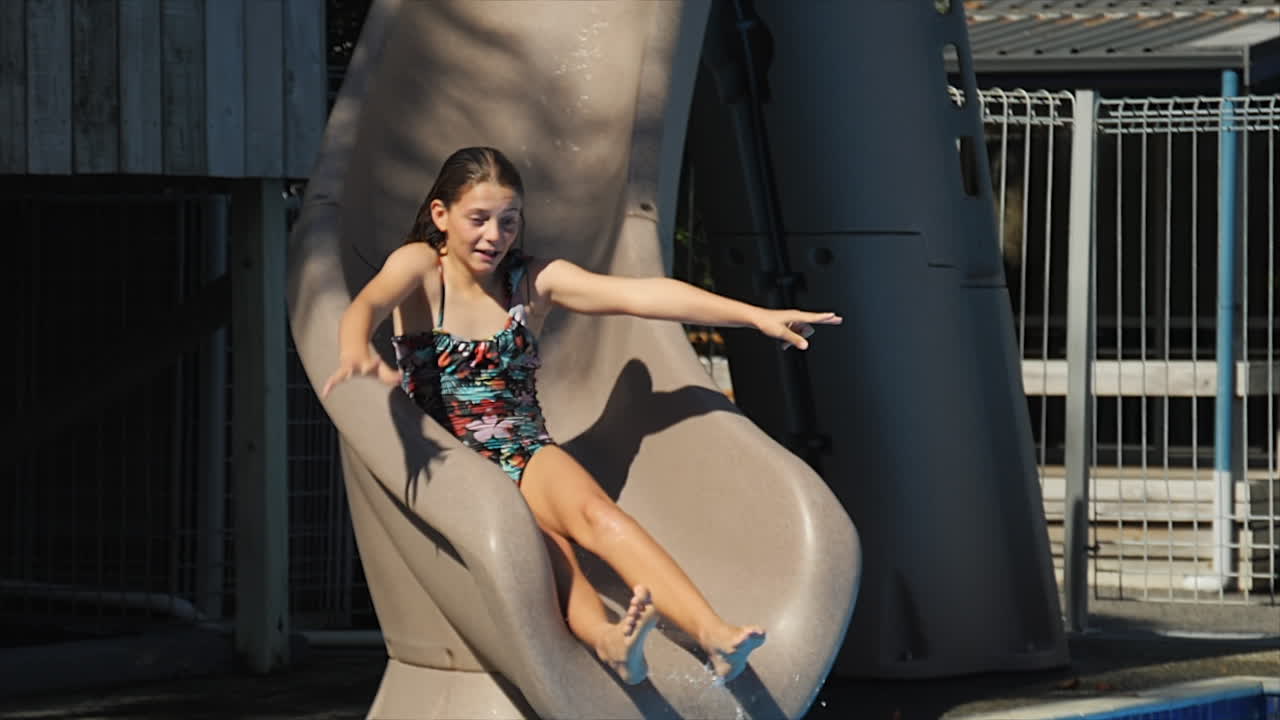 Ragazza preadolescente che posa giocosamente mentre scivola giù da uno scivolo acquatico in una piscina al rallentatore.