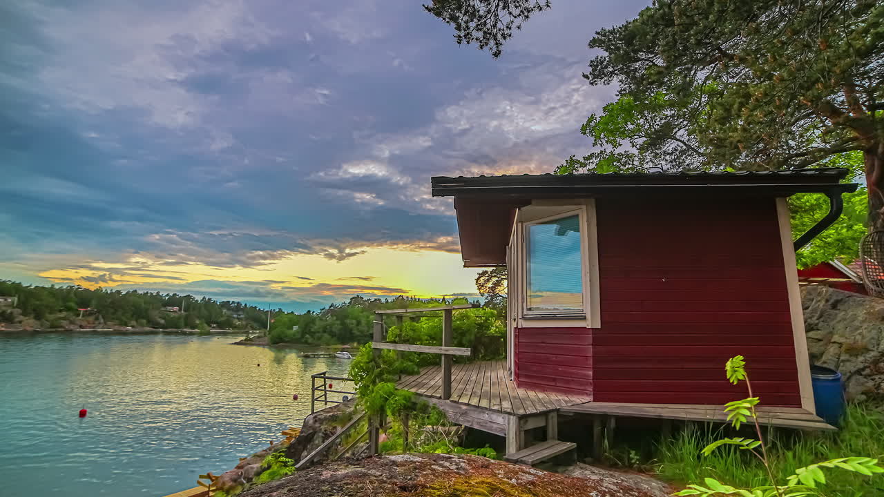 una cabaña roja junto a un lago en suecia al atardecer con una colorida puesta de sol reflejándose en el agua - lapso de tiempo de movimiento