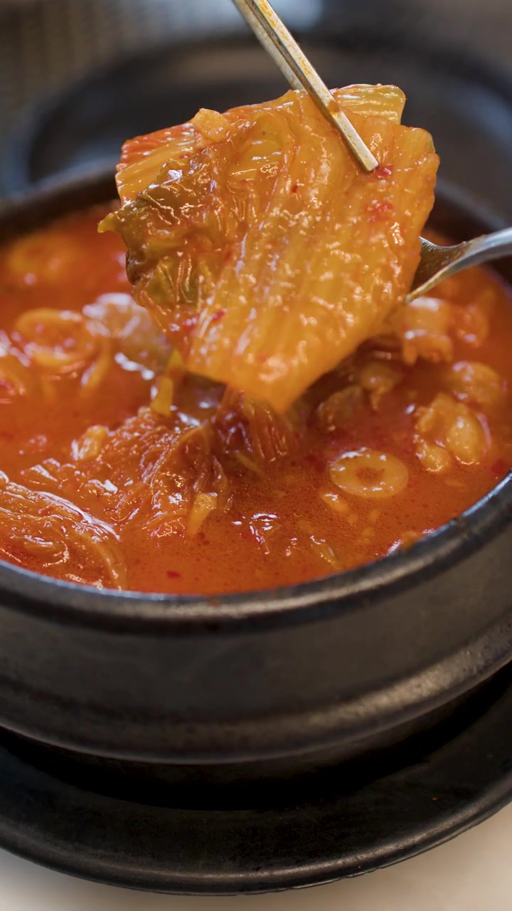 Sumpit logam mengangkat potongan daging babi dan kimchi dari sup tahu merah pedas yang mengepul.