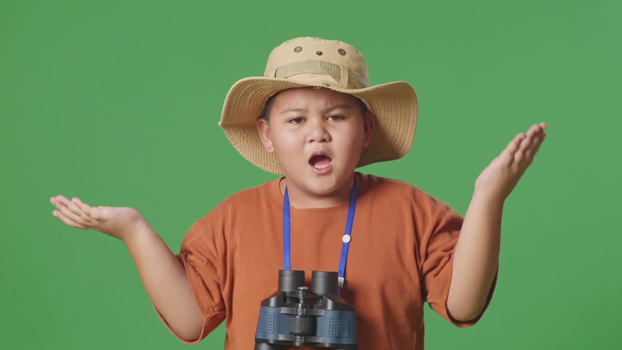 niño turista asiático con un sombrero de pie con dudas y preguntando por qué después de mirar a través de los binoculares. niño investigador examina algo en el fondo de la pantalla verde, concepto de aventura de viaje, de cerca