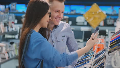 Young-beautiful-married-couple-a-man-and-a-woman-hold-a-smartphone-standing-near-the-showcase-with-smartphones-in-the-appliances-store-choosing-the-best-smart-phone