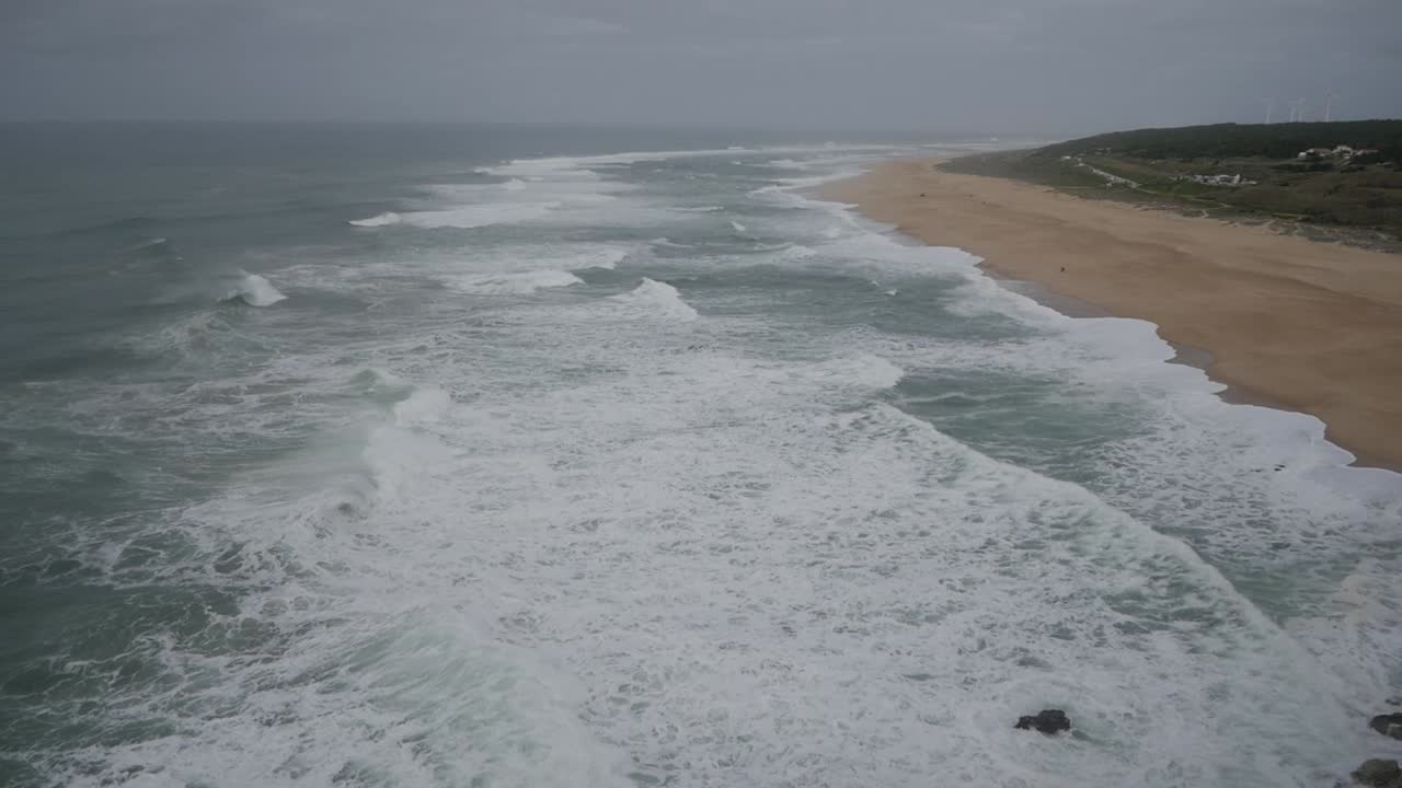 Boundless landscape scenery of Portugal coastline by Atlantic ocean. Praia Do Norte, Nazare. Tide moving waves foam to wild beach. Sand and vegetation. Windmill on distance.
