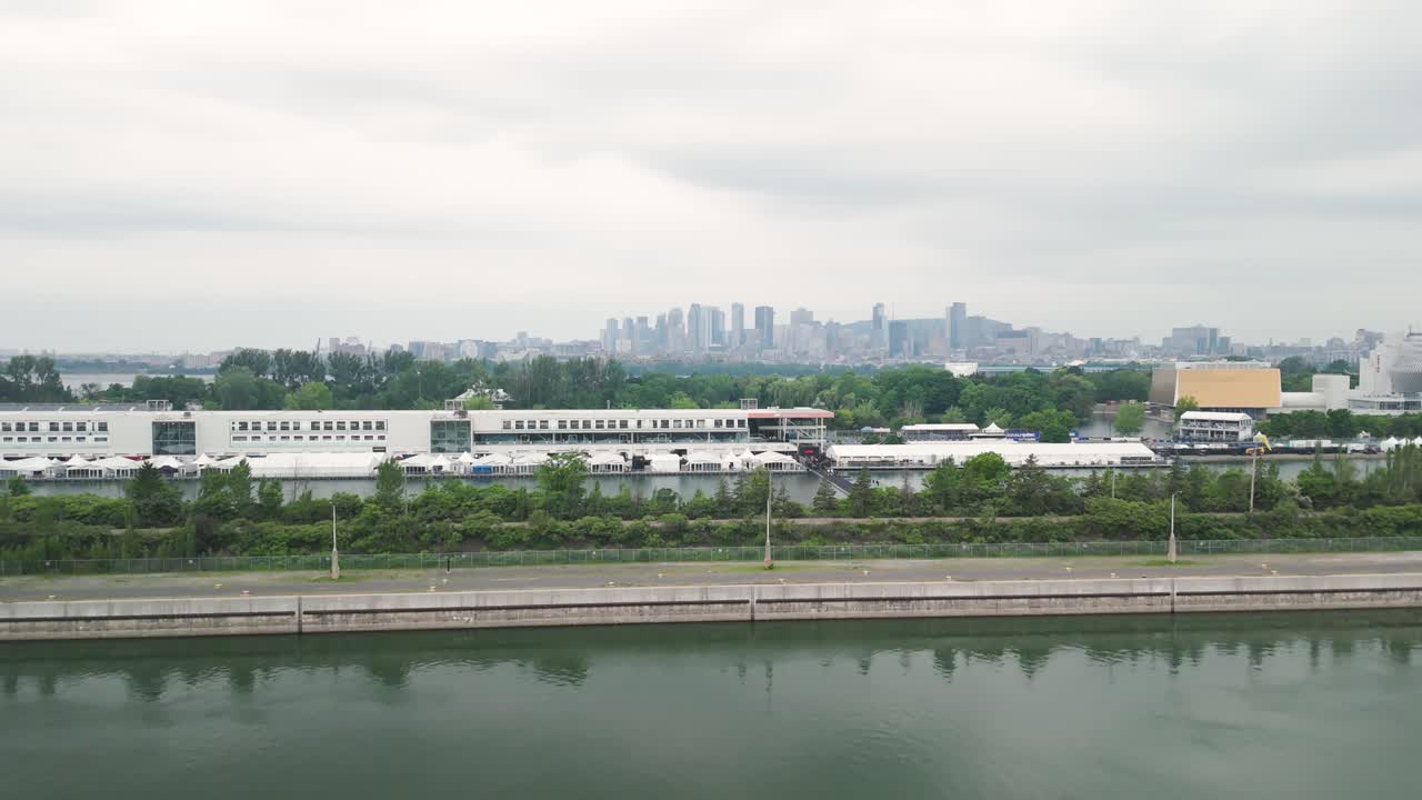Slow aerial drone shot over Montreal’s Circuit Gilles Villeneuve, featuring Espace Paddock, the St. Lawrence River, and the Montreal skyline under an overcast sky.