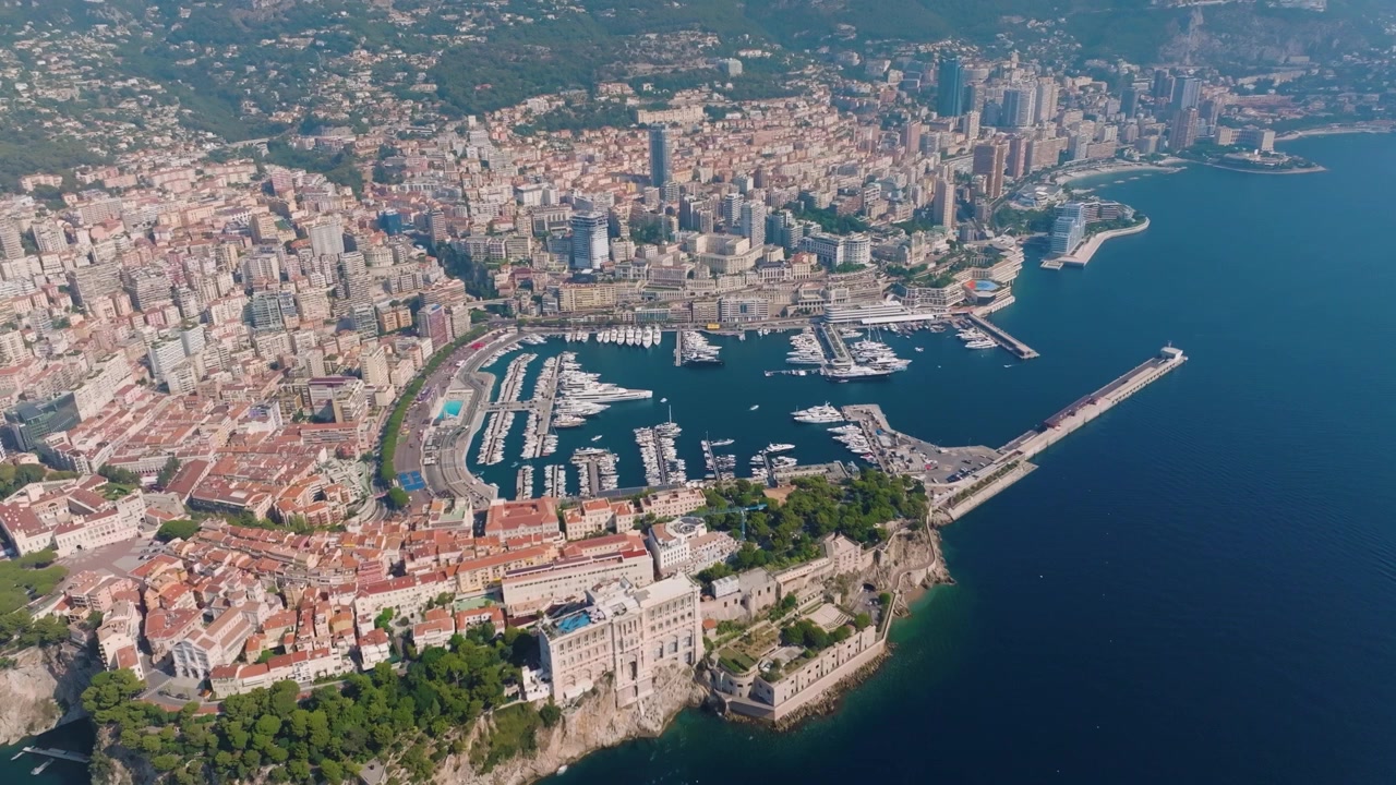 Un filmato cattura lo skyline di Monaco lungo la costa. La panoramica evidenzia edifici, banchine e la disposizione delle strade cittadine. La vista illustra la trafficata area portuale e lo sviluppo urbano.