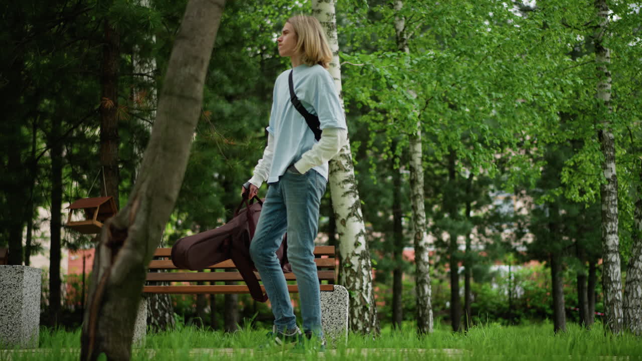 Ung kaukasisk musiker promenerar förbi en bänk, bär en gitarrväska i läder, självsäker gång, soldränkt stig, närmar sig en spelplats, ryggsäck slängd över axeln, avslappnad denim, upplyftande stämning
