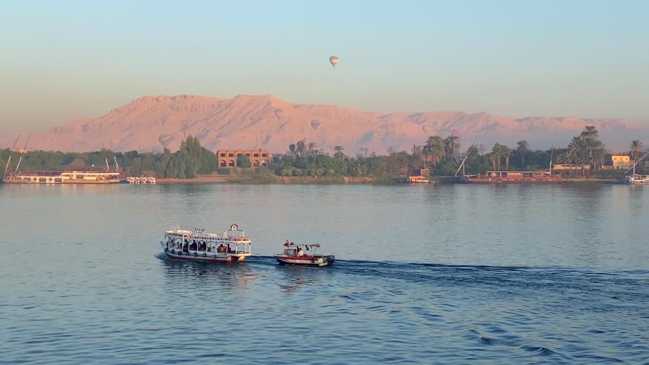 imágenes en 4k de dos pequeños botes navegando en el río nilo frente al valle de los reyes donde varios globos aerostáticos sobrevuelan los cielos de egipto al amanecer