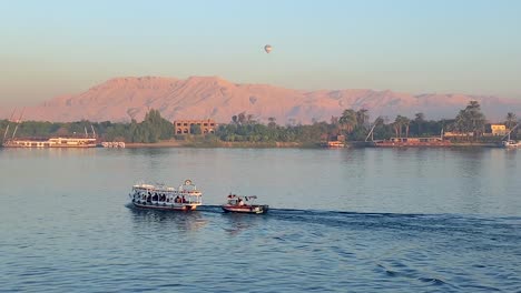 4K-Aufnahmen-Von-Zwei-Kleinen-Booten,-Die-Auf-Dem-Nil-Vor-Dem-Tal-Der-Könige-Segeln,-Wo-Im-Morgengrauen-Mehrere-Heißluftballons-über-Den-Himmel-Ägyptens-Fliegen