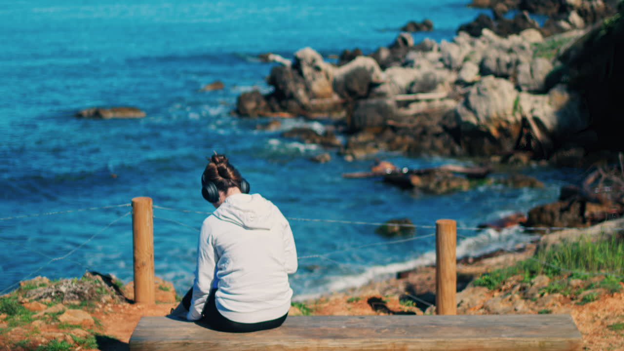 Woman with headphones on her head sitting on a wooden bench with a view of the sea