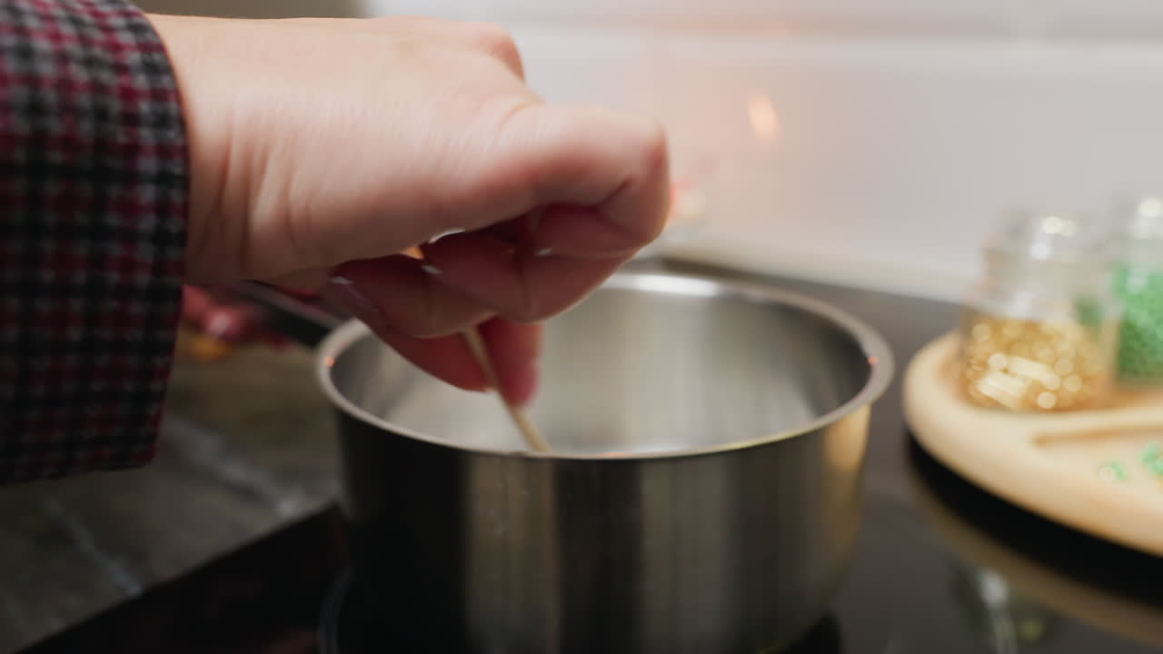 vista de cerca de la mano de una persona que agita el azúcar en una olla con un palo en la estufa mientras que las cuentas coloridas de diferentes tamaños están dispuestas en una bandeja de madera en el fondo
