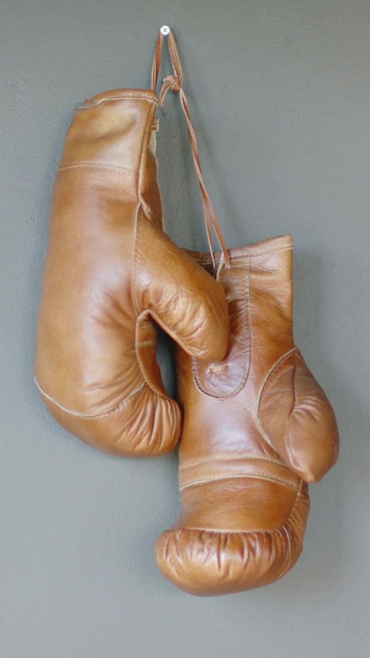 Video verticale: Guanti da boxe in pelle marrone appesi che oscillano su una parete grigia, mostrando lacci e gancio.