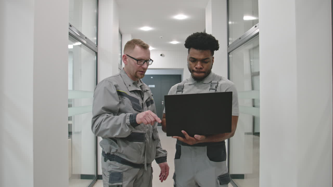 Two technicians or engineers discussing work on a laptop in an office hallway