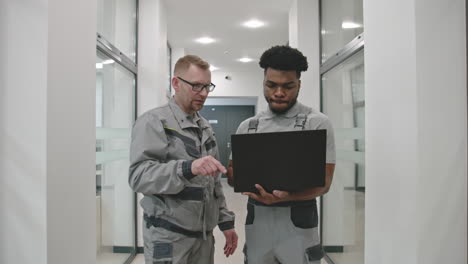 two technicians or engineers discussing work on a laptop in an office hallway