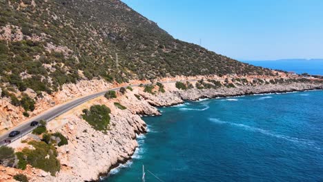 Drone-flying-over-a-mountain-road-with-moving-cars-along-the-coast-over-a-yacht-against-the-backdrop-of-blue-sky-and-blue-water