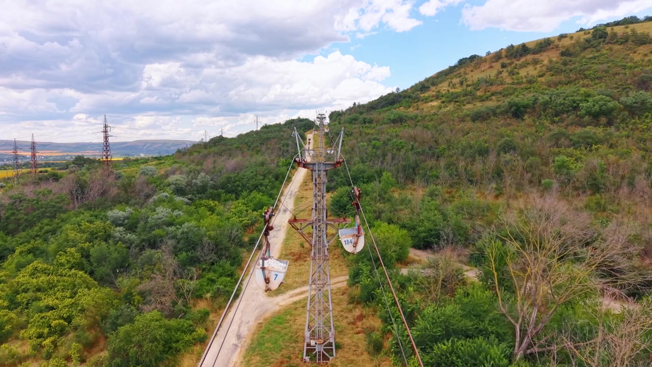 Cableway pylons extending up grassy hillside. Wide aerial view shows industrial steel towers and cable lines above countryside path.