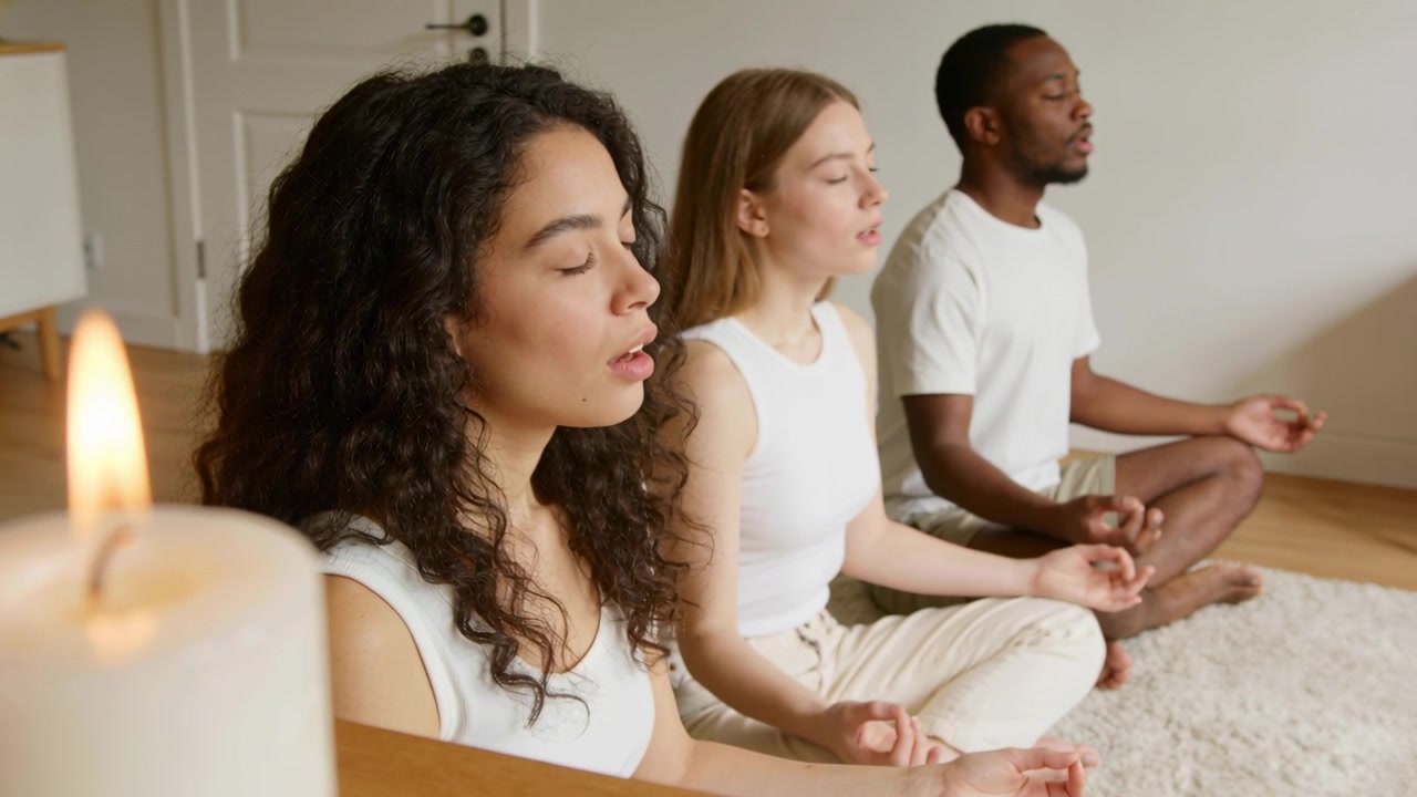 La telecamera rivela tre adulti seduti che meditano a casa, indossando magliette bianche, con una candela a colonna accesa.
