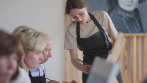 a female teacher shows a retired man how to draw a picture with paints and a brush at courses for the elderly. a senior man draws a picture to a group of pensioners