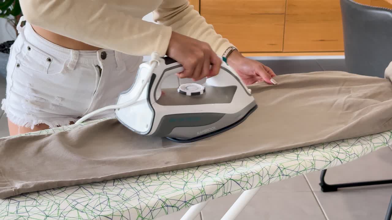 A young woman in casual clothing uses a steam iron to press beige pants on a patterned ironing board in a bright, contemporary home setting