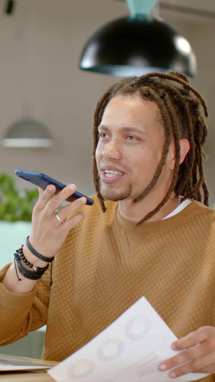 Video verticale: Uomo in maglione che tiene un telefono blu alla bocca e fornisce un aggiornamento su un grafico al tavolo.