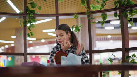 focused young woman in plaid shirt applying makeup on jaw and cheeks while using mirror, various beauty products and personal items arranged on wooden table