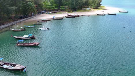 drone footage captures longtail boats along sarasin beach, phuket. clear waters, sandy shores, and lush greenery create a serene coastal scene