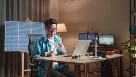 asian man sitting in front of solar cell looks at wind turbine while typing on a laptop at the office