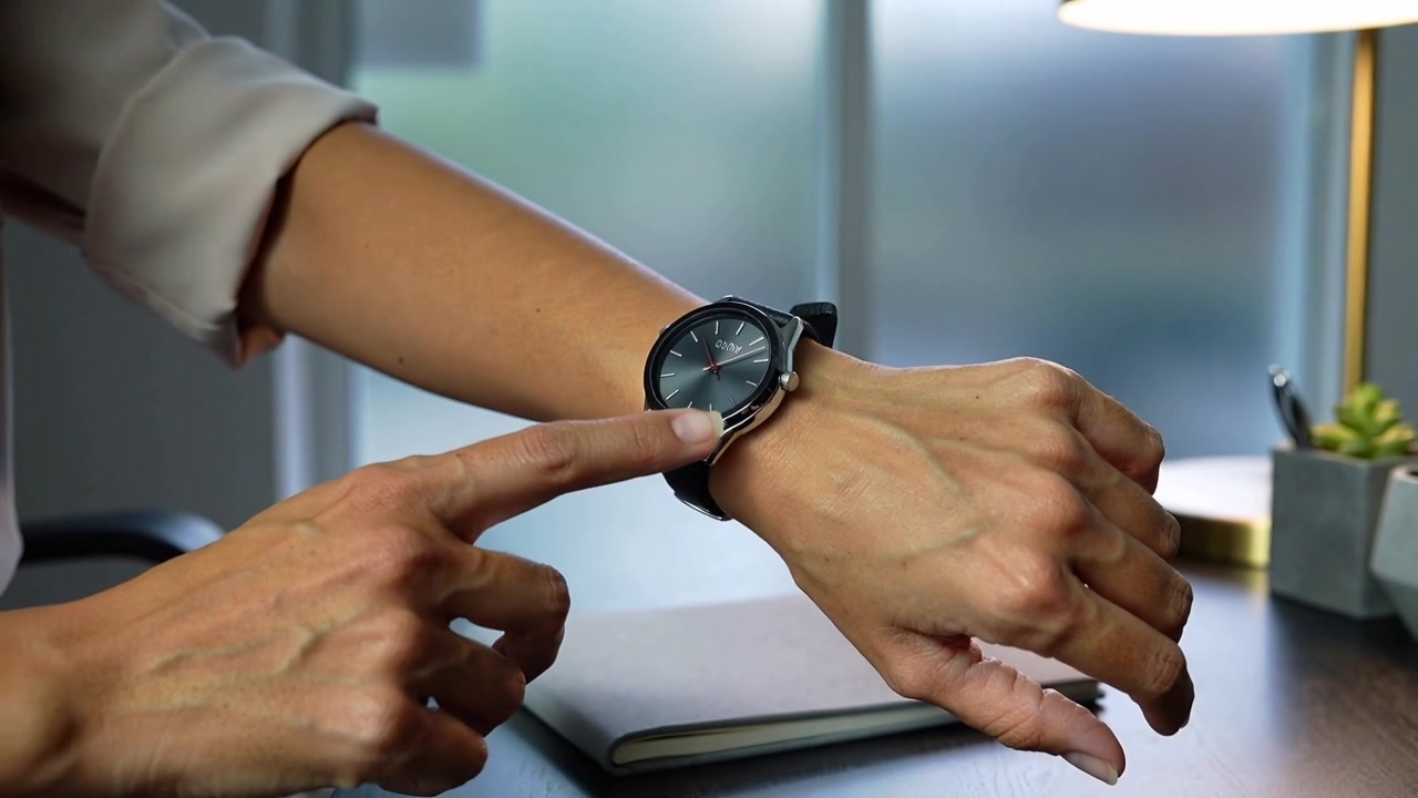 Tapping right finger, woman wearing rolled sleeves adjusting wristwatch at office desk for fit