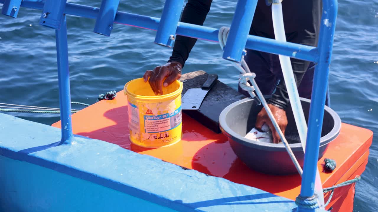 A person fillets fish on a boat in Phuket, Thailand. Bright sunlight, vibrant colors, and ocean waves create a lively atmosphere