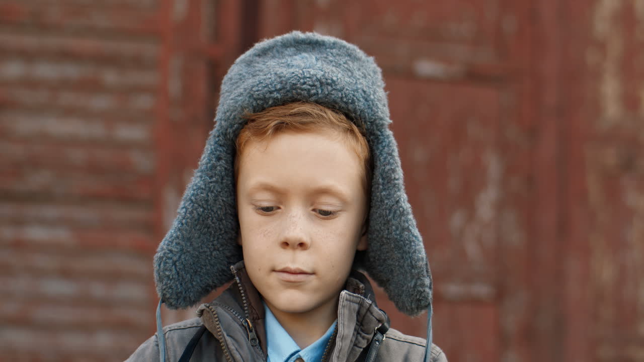 портрет милого рыжеволосого мальчика в шляпе-авиаторе, фотографирующегося с винтажной камерой на открытом воздухе на фоне деревянной стены