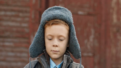 portrait of a cute little red haired boy in aviator hat taking photos with vintage camera outdoors on a wooden wall background