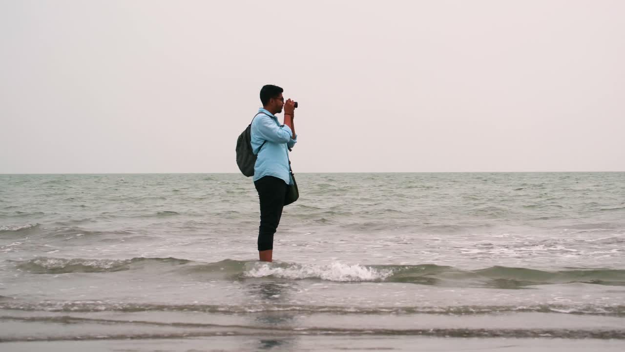 Male photographer standing in shallow water of the shore facing side view with pants curled up to avoid the waves taking photos