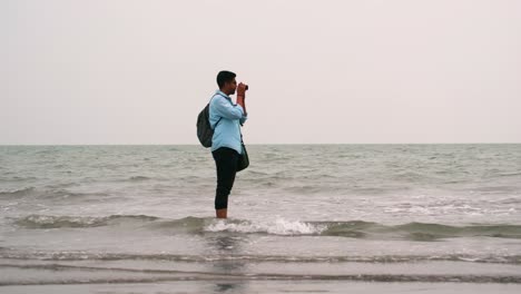 Ein-Männlicher-Fotograf-Steht-Im-Seichten-Wasser-Des-Ufers-Mit-Blick-Auf-Die-Seitenansicht-Und-Hat-Die-Hosen-Zusammengerollt,-Um-Den-Wellen-Beim-Fotografieren-Auszuweichen