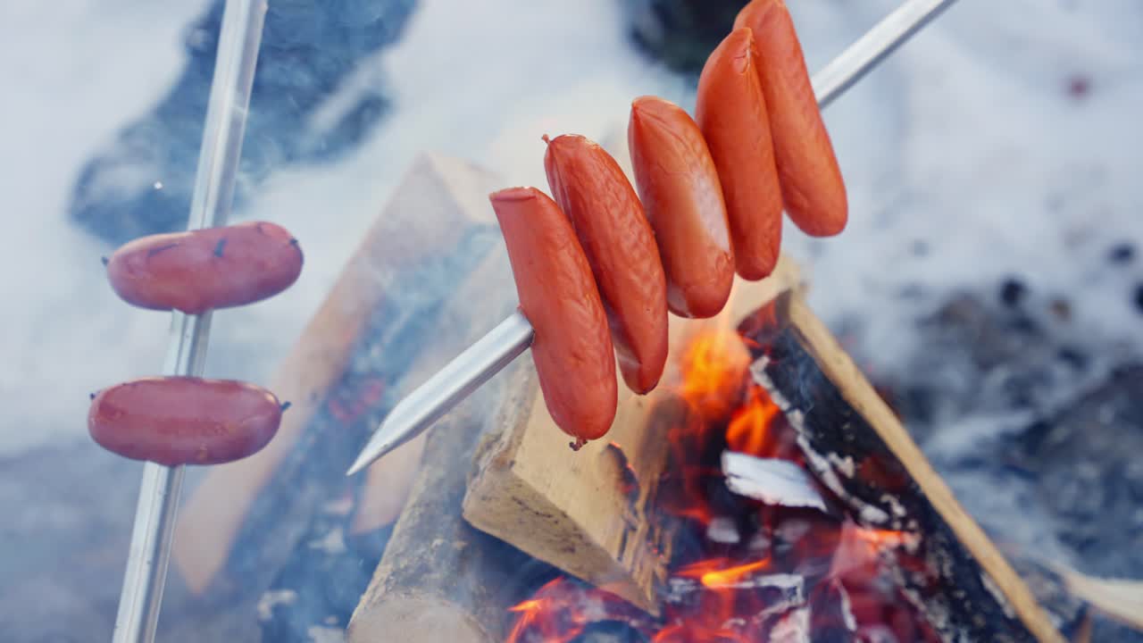 Salsicce che cuociono sopra una fiamma libera mentre la legna brucia, creando una scena rustica di barbecue all'aperto, infilzate, al rallentatore